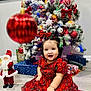 toddler, child, red_dress, christmas_tree, holiday, decorations, santa_figurine, ornament, smiling, indoors, festive, presents, floor, curly_hair, plaid, joyful, celebration, seasonal, cute, portrait