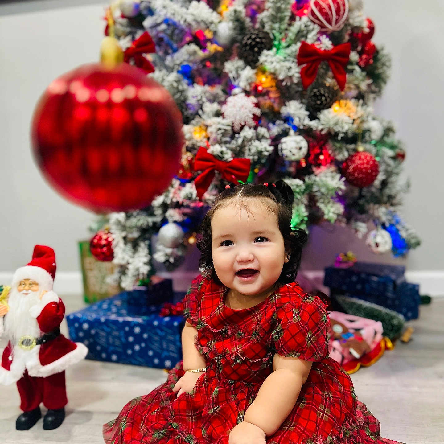Bianca Castellanos is registered to the contest to win money with this photo: celebration, child, christmas_tree, curly_hair, cute, decorations, festive, floor, holiday, indoors, joyful, ornament, plaid, portrait, presents, red_dress, santa_figurine, seasonal, smiling, toddler
