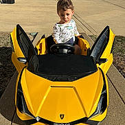 Langston is registered to the contest to win money with this photo: child, toddler, toy_car, yellow_car, outdoor, driveway, sunny, sidewalk, concrete, small_vehicle, playtime, cute, young_child, car_doors, steering_wheel, casual_clothing, greenery, residential_area, daylight, expression
