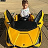 child, toddler, toy_car, yellow_car, outdoor, driveway, sunny, sidewalk, concrete, small_vehicle, playtime, cute, young_child, car_doors, steering_wheel, casual_clothing, greenery, residential_area, daylight, expression
