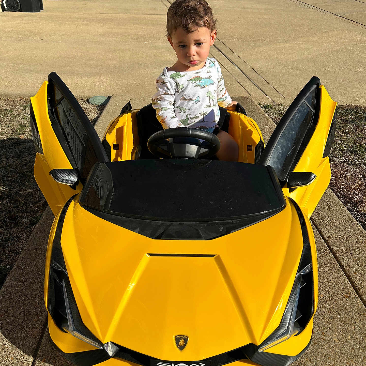 Langston is registered to the contest to win money with this photo: child, toddler, toy_car, yellow_car, outdoor, driveway, sunny, sidewalk, concrete, small_vehicle, playtime, cute, young_child, car_doors, steering_wheel, casual_clothing, greenery, residential_area, daylight, expression