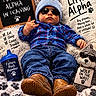 baby, child, infant, portrait, sunglasses, beanie, plaid_shirt, blue_clothing, jeans, boots, blanket, teddy_bear, sippy_cup, indoor, lying_down, smiling, fashion, text_overlay, props, playful