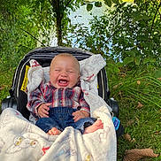 Vincent is registered to the contest to win money with this photo: baby, infant, child, laughing, smiling, stroller, blanket, outdoor, nature, greenery, water, lake, forest, grass, plaid_shirt, jeans, portrait, seated, sunlight, happy
