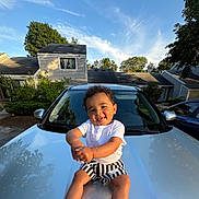 Kaizen is registered to the contest to win money with this photo: toddler, child, car, hood, smiling, playful, outdoor, daytime, sky, clouds, house, suburban, curly_hair, white_shirt, striped_shorts, happy, sunlight, reflection, nature, greenery