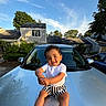toddler, child, car, hood, smiling, playful, outdoor, daytime, sky, clouds, house, suburban, curly_hair, white_shirt, striped_shorts, happy, sunlight, reflection, nature, greenery