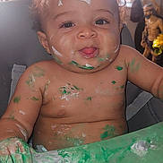 Legend is registered to the contest to win money with this photo: baby, child, paint, messy, smiling, face, arms, table, highchair, curly_hair, indoor, skin, green_paint, white_paint, mischievous, expression, portrait, happy, playful, fun