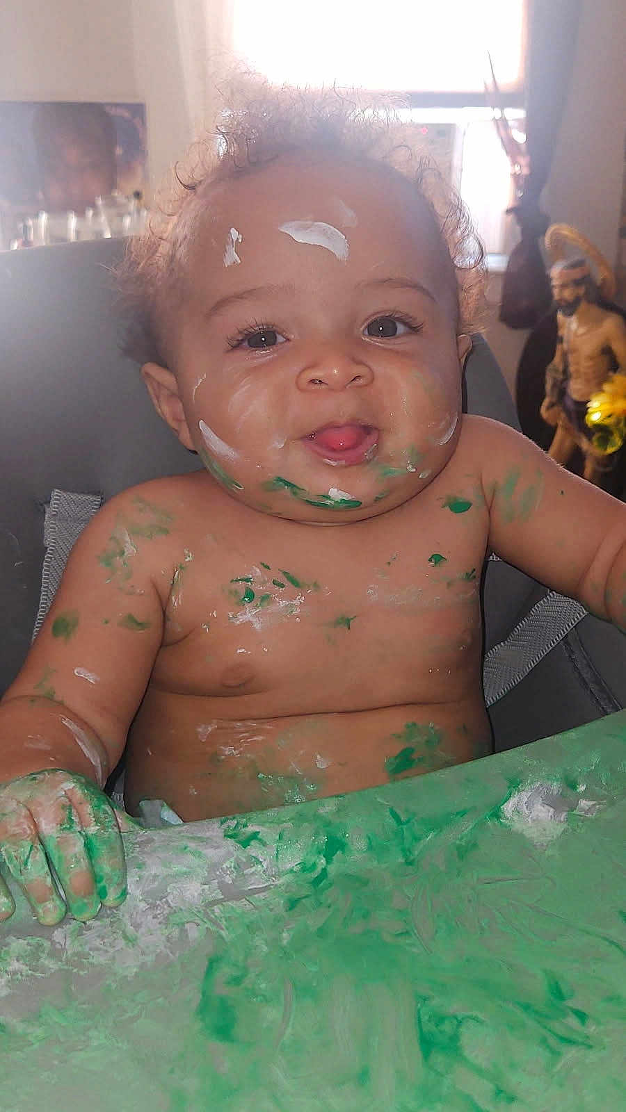 Legend is registered to the contest to win money with this photo: baby, child, paint, messy, smiling, face, arms, table, highchair, curly_hair, indoor, skin, green_paint, white_paint, mischievous, expression, portrait, happy, playful, fun