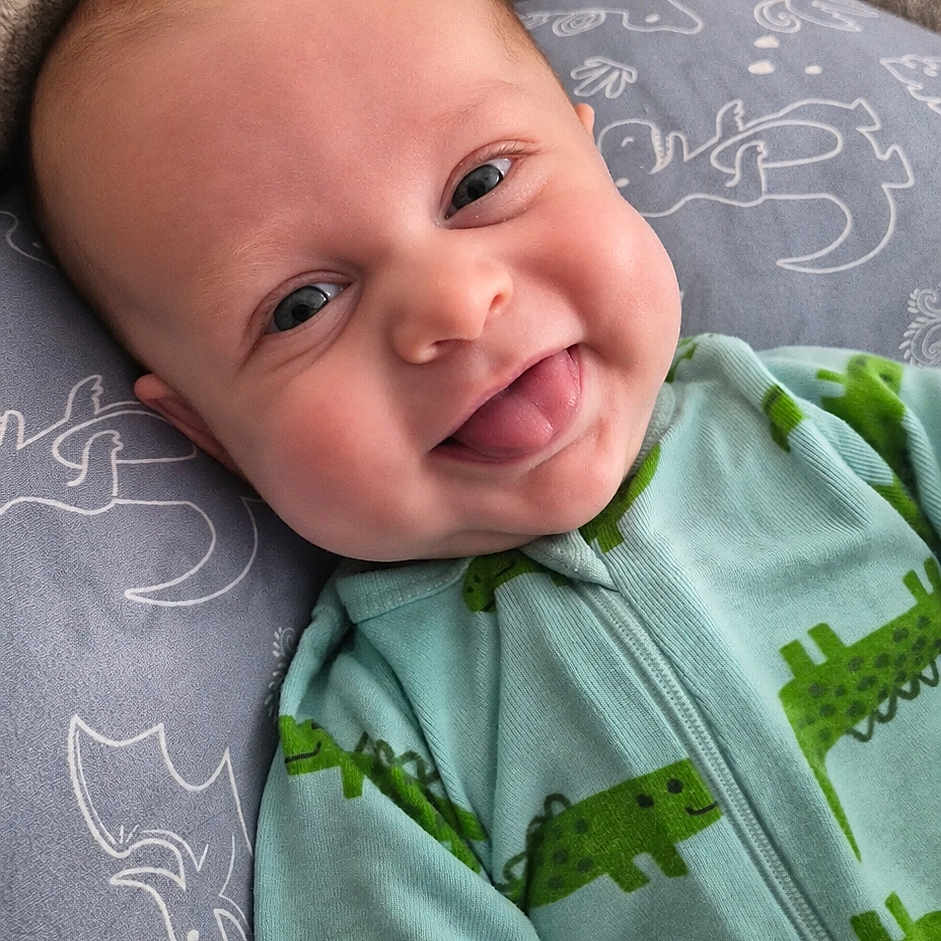 Charlie joined the competition — help win amazing prizes! baby, infant, child, smiling, tongue_out, portrait, close_up, indoor, lying_down, blanket, pillow, dinosaur_clothing, green_outfit, cozy, happy, face, eyes, head, soft_lighting, photography