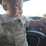 Gunner is registered to the contest to win money with this photo: baby, child, infant, overalls, dinosaur_print, white_shirt, steering_wheel, car_interior, dashboard, dodge, hand, face, cheeks, curious, sitting, vehicle, window, daylight, cute, portrait