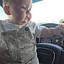 baby, child, infant, overalls, dinosaur_print, white_shirt, steering_wheel, car_interior, dashboard, dodge, hand, face, cheeks, curious, sitting, vehicle, window, daylight, cute, portrait