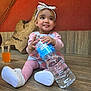 baby, bow, child, cute, floor, girl, glass, headband, indoor, infant, orange_juice, pink_clothing, playful, shoes, sitting, smiling, tongue_out, wall, water_bottle, wooden_floor
