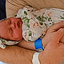 newborn, baby, swaddled, floral_blanket, headband, arms, hospital_wristband, resting, sleeping, person, skin, hand, comfort, parent, tenderness, closeup, indoor, human, infant, peaceful