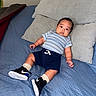 baby, bed, blue_bedspread, casual_clothing, child, cute, furniture, home, indoor, infant, looking_at_camera, navy_shorts, person, pillow, portrait, relaxed, resting, sneakers, socks, striped_shirt