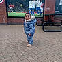 toddler, child, snowsuit, blue_jacket, brick_pavement, outdoor, soccer_poster, sports, curious, standing, pavement, park_bench, table, casual, young_child, winter_clothing, urban, daylight, public_space, expression