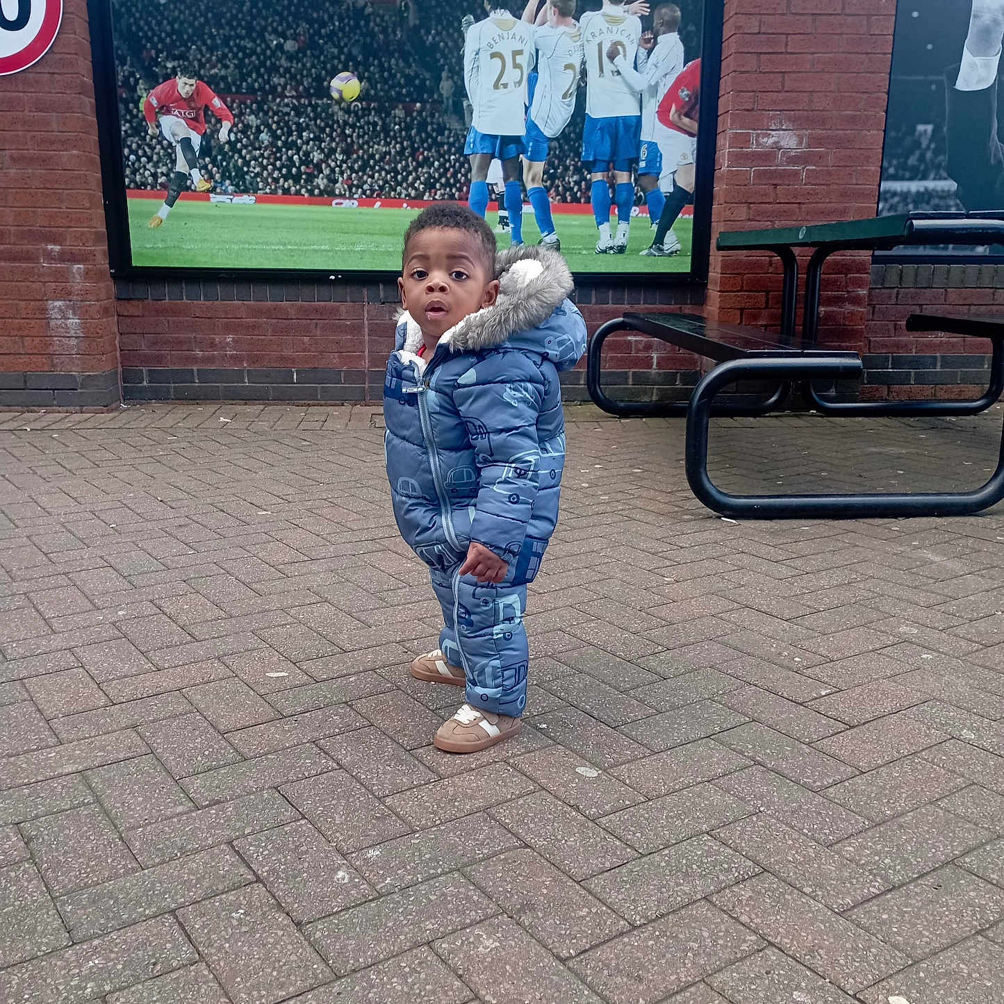 Divine Ntwayagae is registered to the contest to win money with this photo: blue_jacket, brick_pavement, casual, child, curious, daylight, expression, outdoor, park_bench, pavement, public_space, snowsuit, soccer_poster, sports, standing, table, toddler, urban, winter_clothing, young_child