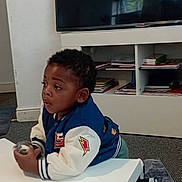 Divine Ntwayagae is registered to the contest to win money with this photo: black_hair, books, boy, carpet, casual_clothing, child, curious, expression, floor, furniture, holding, indoor, jacket, leaning, room, shelf, sneakers, table, television, young
