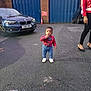 toddler, child, parking_lot, car, bmw, blue_garage_door, brick_wall, woman, sidewalk, asphalt, casual_clothing, red_shirt, jeans, white_shoes, outdoor, urban, curious, standing, person, daytime