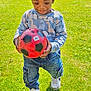 child, boy, hat, soccer_ball, grass, outdoor, sunlight, jeans, sneakers, playing, dinosaur_sweater, happy, nature, greenery, casual_clothing, daylight, portrait, smiling, young_child, sports