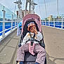 child, stroller, bridge, urban, sidewalk, cap, sunglasses, shoes, clothing, outdoor, person, hand, road, railing, sky, building, casual, baby, transport, relaxed