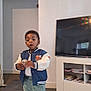 Divine Ntwayagae is registered to the contest to win money with this photo: ball, bookshelf, boy, carpet, child, curious, expression, furniture, holding, indoor, jacket, room, shorts, sneakers, standing, table, television, toddler, white_wall