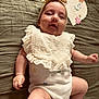baby, infant, headband, bow, bib, lying_down, bedspread, soft_texture, cute, chubby_cheeks, smiling, clothing, skin, hand, foot, decor, flower, portrait, child, indoors