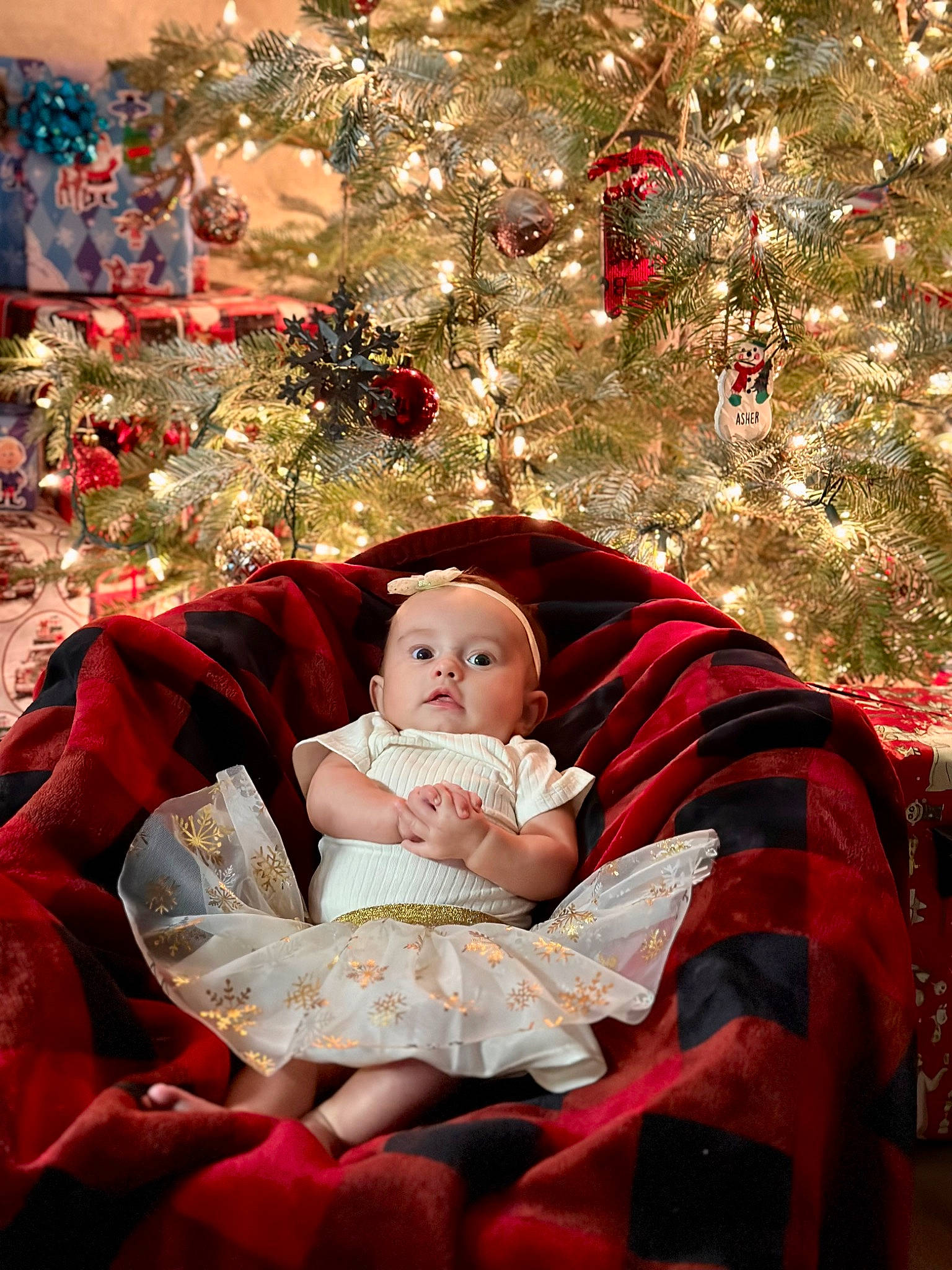 Rhea is registered to the contest to win money with this photo: baby, carmine, christmas, christmas_decoration, christmas_lights, christmas_ornament, christmas_tree, comfort, conifer, dress, event, happy, holiday, holiday_ornament, interior_design, ornament, person, sitting, toddler, tradition