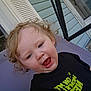 child, toddler, curly_hair, blue_eyes, black_shirt, text_on_clothing, outdoor, porch, door, house_siding, purple_chair, expression, happy, smiling, person, clothing, young_child, cute, casual, daylight