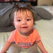 Evelyn joined the competition — help win amazing prizes! toddler, child, girl, pigtails, orange_shirt, gray_pants, bed, remote_control, lotion_bottle, indoors, cute, smiling, drool, young_child, portrait, person, sitting, hair, face, eyes