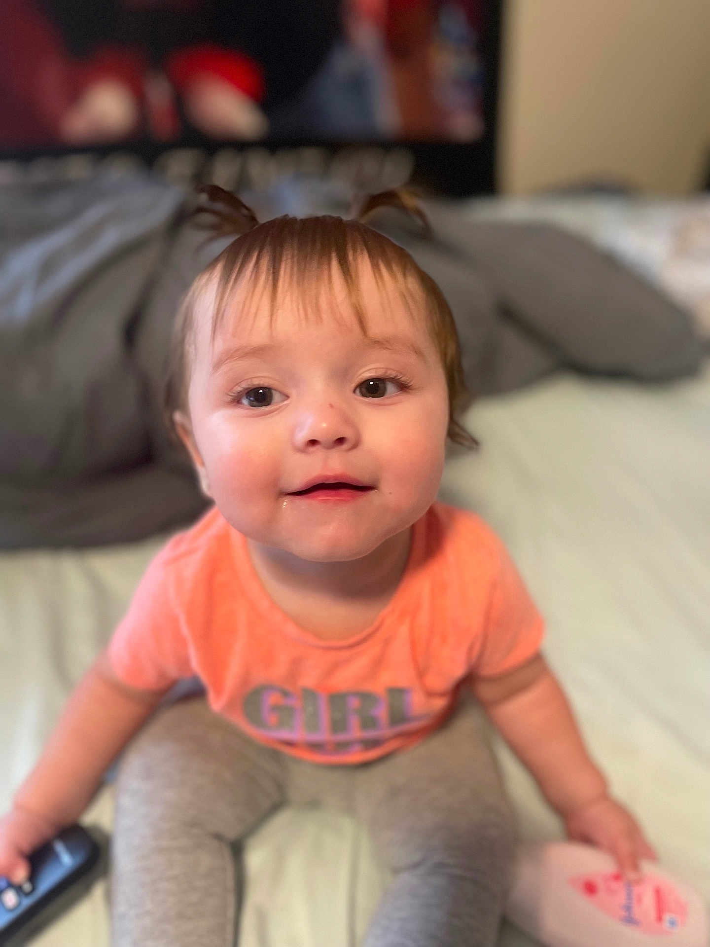 Evelyn joined the competition — help win amazing prizes! toddler, child, girl, pigtails, orange_shirt, gray_pants, bed, remote_control, lotion_bottle, indoors, cute, smiling, drool, young_child, portrait, person, sitting, hair, face, eyes
