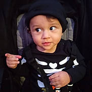 Indigo is registered to the contest to win money with this photo: child, toddler, baby, face, black_clothing, hat, skeleton_outfit, stroller, safety_harness, pointing, curious, indoor, portrait, young_child, cute, expression, seat, black_background, person, closeup