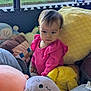 toddler, child, plush_toys, pink_clothing, cushions, indoor, window, soft_light, yellow_pillow, stuffed_animal, cute, portrait, cozy, seated, curious, baby, play_area, colorful, soft_texture, young_child