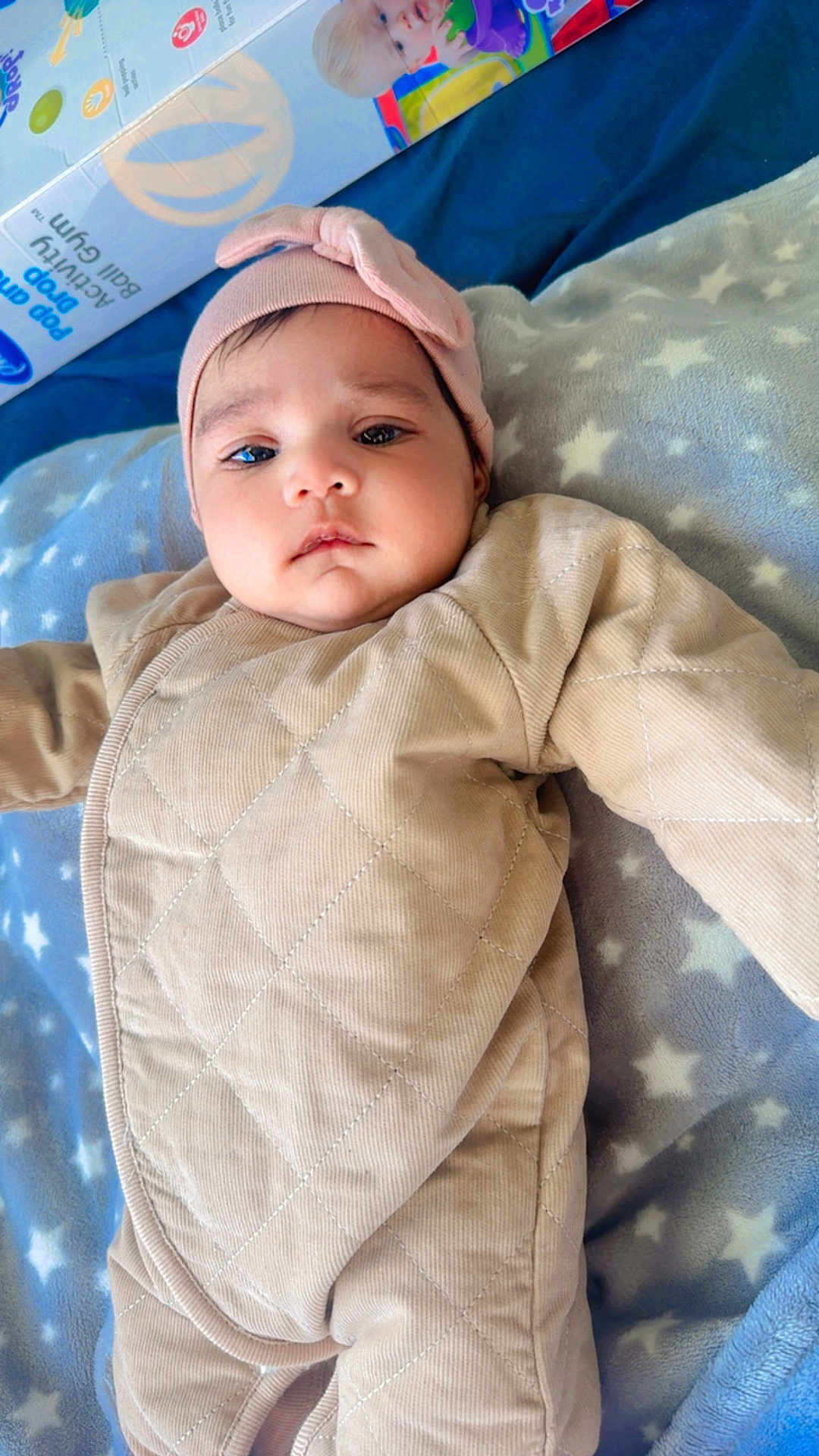 Sabreen is registered to the contest to win money with this photo: baby, infant, blanket, headband, quilted_clothing, star_pattern, blue, beige, pink, face, portrait, child, lying_down, soft_texture, indoor, box, toy_box, curious, cute, young