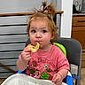 Lizzy is registered to the contest to win money with this photo: toddler, child, high_chair, snack, pink_shirt, curly_hair, hair_tie, indoor, kitchen, eating, food, curious, seat, table, person, baby, cute, face, expression, young_child