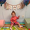 toddler, child, toy, balloon, play_mat, pink_clothing, socks, wall_decoration, banner, indoor, smiling, tongue_out, colorful, floor_mat, animal_toy, plastic_toy, celebration, cute, person, happy