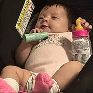 Skye is registered to the contest to win money with this photo: baby, infant, car_seat, toy, baby_bottle, pink_socks, onesie, child, cute, smiling, indoor, seatbelt, warning_label, hands, legs, face, clothing, holding, small_toy, happy