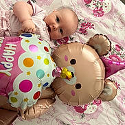 Milaya a rejoint le concours — aidez-le/la à gagner de superbes lots ! baby, infant, balloon, party, pink_clothing, floral_bedspread, toy, celebration, cute, lying_down, face, child, indoor, decor, soft_lighting, head, hand, smiling, birthday, party_hat