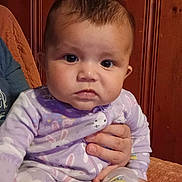 Memory is registered to the contest to win money with this photo: baby, child, infant, portrait, sitting, serious_expression, wide_eyes, brown_hair, pajamas, purple_clothing, patterned_clothing, adult_hand, support, blanket, wooden_background, indoor, close_up, cozy, home, photography