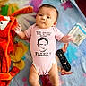 baby, infant, child, lying_down, portrait, face, smiling, pink_onesie, clothing, blanket, colorful, plush_toy, red_toy, remote_control, bed, indoor, cozy, playful, baby_supplies, text_on_clothing