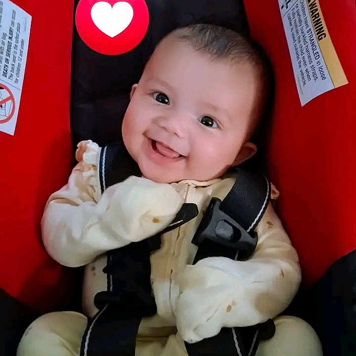 Memory is registered to the contest to win money with this photo: baby, child, infant, smiling, happy, car_seat, safety_harness, portrait, indoor, close_up, face, eyes, mouth, mitten, onesie, red, black, heart_icon, seated, automobile