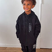 Kessim participe au concours pour gagner de l'argent avec cette photo : child, smiling, standing, indoor, black_jacket, under_armour, black_pants, colorful_sneakers, curly_hair, headband, wooden_floor, white_wall, door, happy, person, fashion, casual, portrait, full_body, clothing
