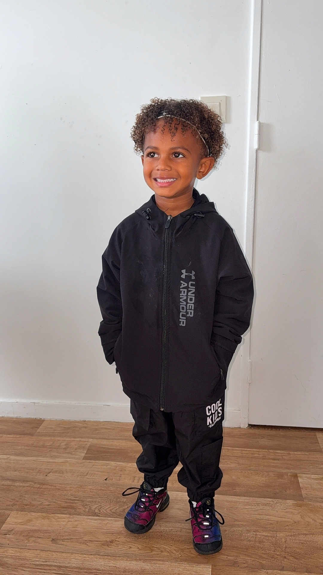 Kessim participe au concours pour gagner de l'argent avec cette photo : child, smiling, standing, indoor, black_jacket, under_armour, black_pants, colorful_sneakers, curly_hair, headband, wooden_floor, white_wall, door, happy, person, fashion, casual, portrait, full_body, clothing