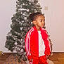 boy, carpet, child, christmas_tree, cozy, decoration, festive, gift_wrap, hardwood_floor, holiday, indoor, looking_sideways, ornament, pine_tree, portrait, present, red_tracksuit, silver_bauble, standing, white_shoes
