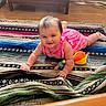 baby, child, pink_dress, toy, rug, floor, wooden_cabinet, indoor, smiling, happy, crawling, playful, colorful, sock, person, home, furniture, head, face, bright
