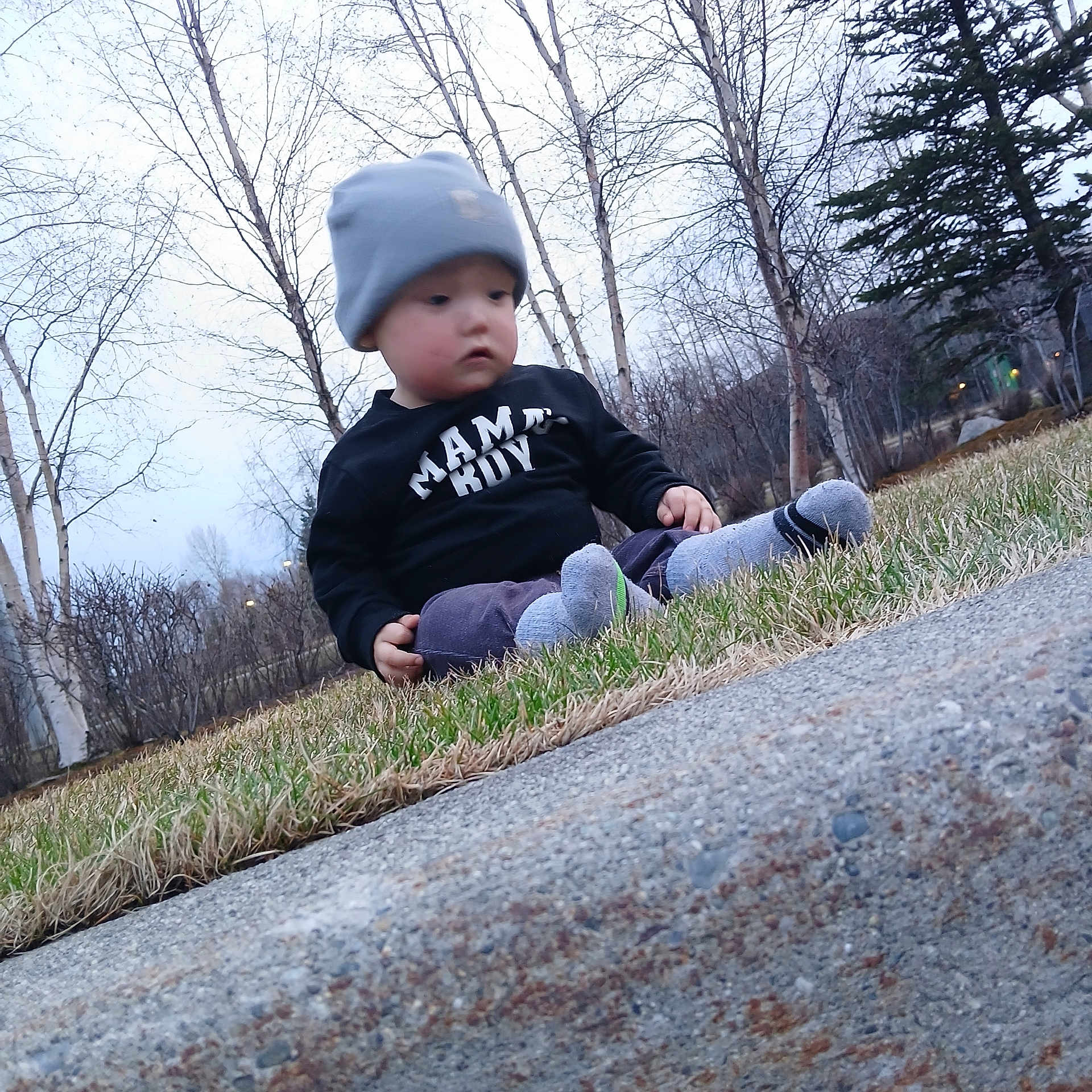 Jonathan is registered to the contest to win money with this photo: toddler, child, grass, beanie, socks, concrete, outdoor, trees, winter, curious, clothing, nature, sitting, side_view, young_child, casual, daylight, park, bare_trees, cold_weather