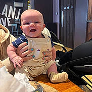 Parker joined the competition — help win amazing prizes! baby, smile, overalls, striped_shirt, hands, nail_polish, table, food, knife, indoor, wooden_furniture, high_chair, person, face, child, happy, cute, seated, cozy, restaurant