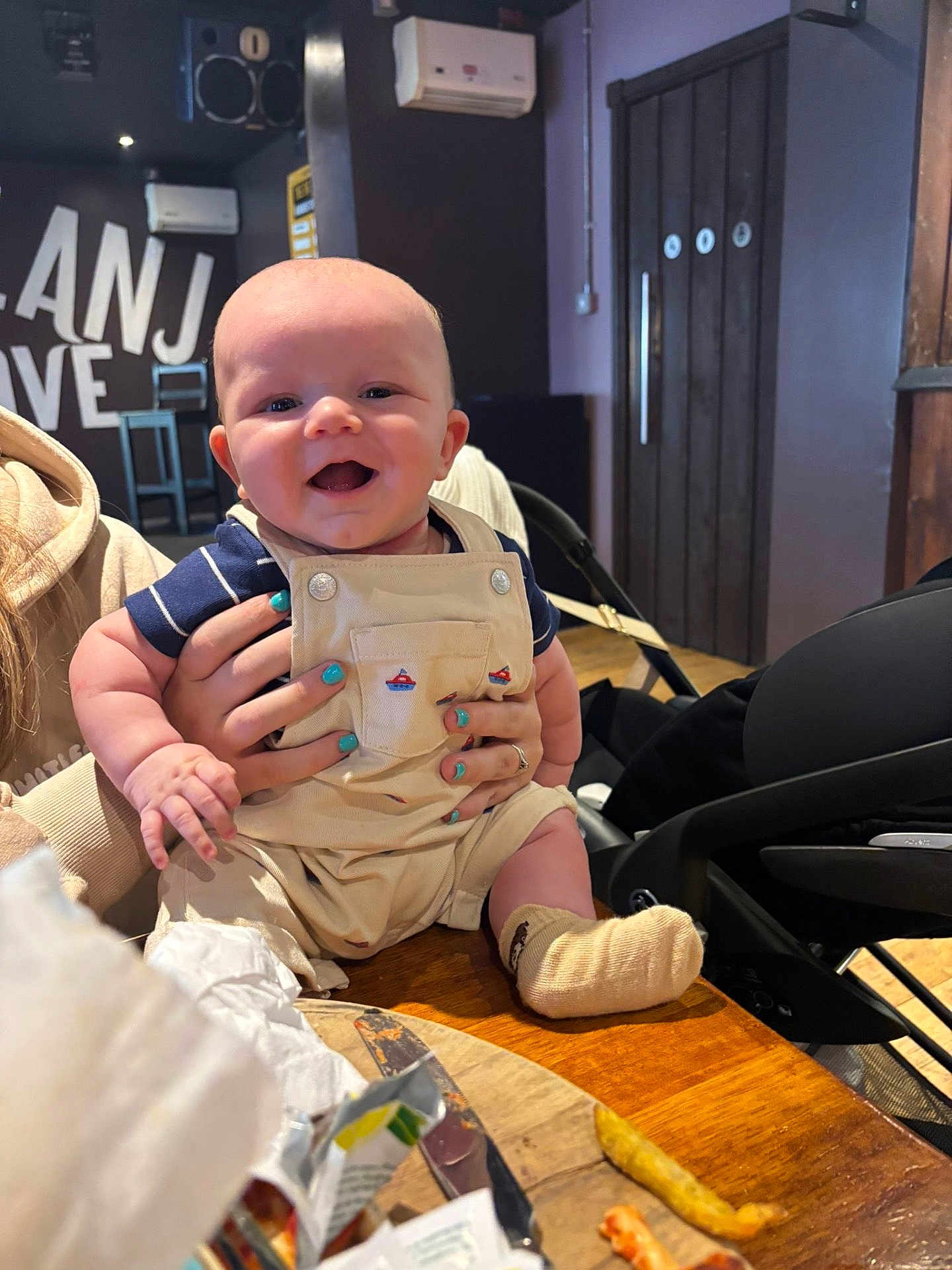 Parker joined the competition — help win amazing prizes! baby, smile, overalls, striped_shirt, hands, nail_polish, table, food, knife, indoor, wooden_furniture, high_chair, person, face, child, happy, cute, seated, cozy, restaurant