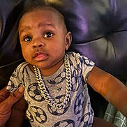 Zhakari is registered to the contest to win money with this photo: baby, child, onesie, soccer_ball_pattern, necklace, chain, leather_couch, hand, indoor, curious_expression, person, portrait, infant, clothing, sitting, closeup, skin, face, casual, cute