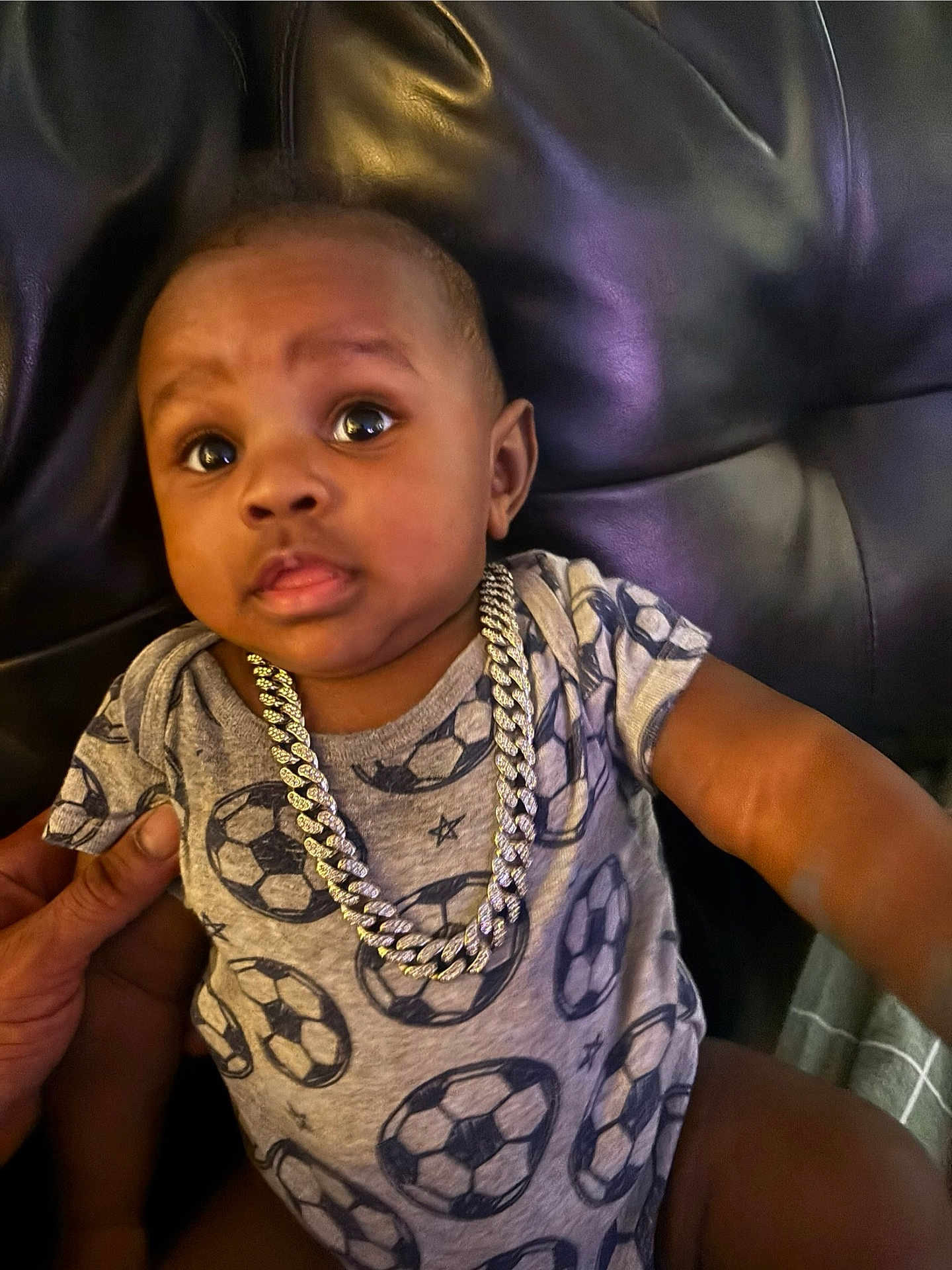 Zhakari is registered to the contest to win money with this photo: baby, child, onesie, soccer_ball_pattern, necklace, chain, leather_couch, hand, indoor, curious_expression, person, portrait, infant, clothing, sitting, closeup, skin, face, casual, cute
