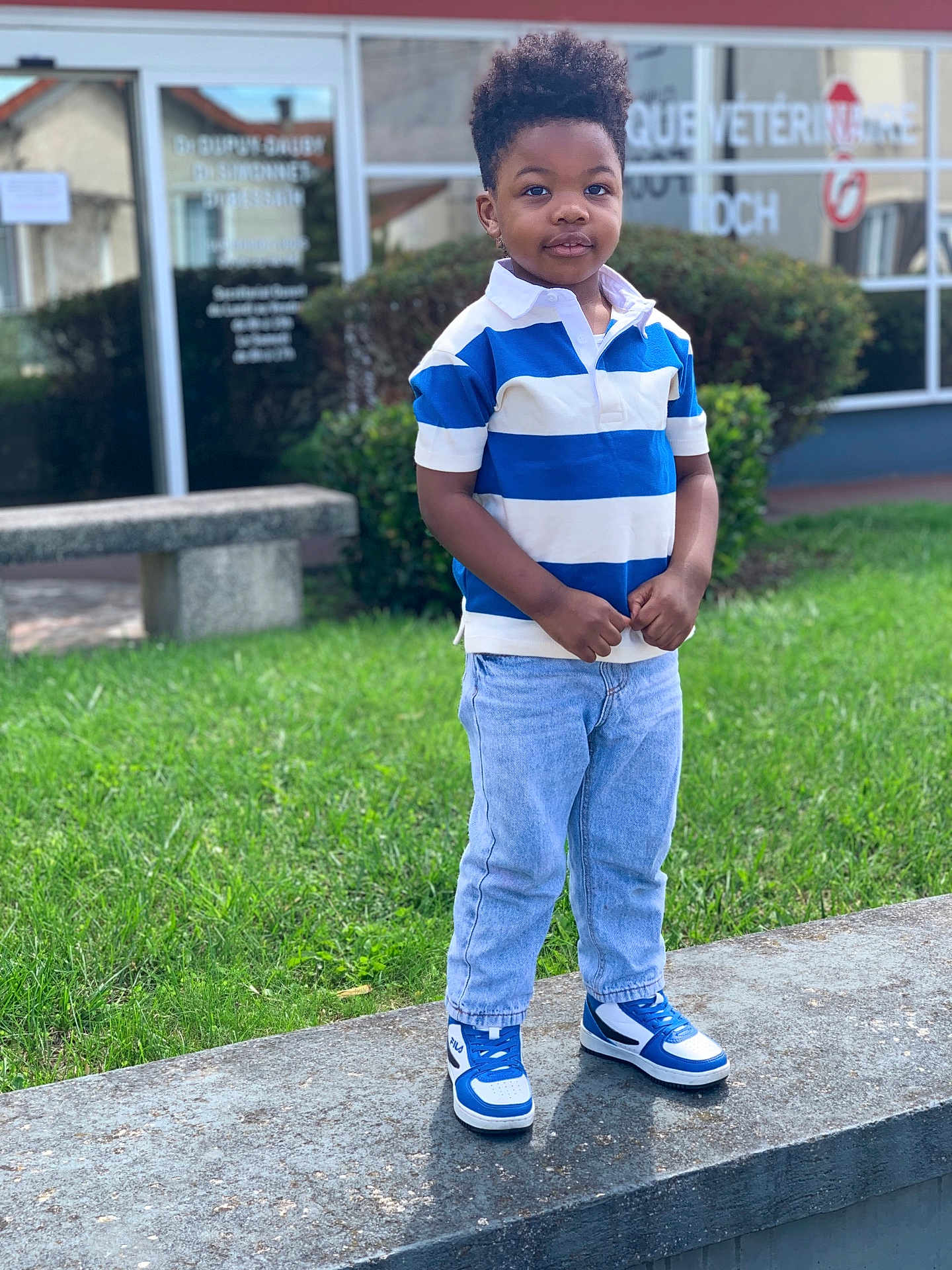Enzo-Ebuwa Omosumwen a rejoint le concours — aidez-le/la à gagner de superbes lots ! child, boy, standing, outdoor, grass, jeans, polo_shirt, sneakers, blue, white, building, window, bench, greenery, casual_clothing, portrait, smiling, cute, fashion, urban