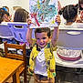 child, boy, painting, art_class, colorful, smiling, happy, indoor, wooden_table, chairs, people, kids, creative, drawing, playful, shirt, shorts, floor, hair, expression